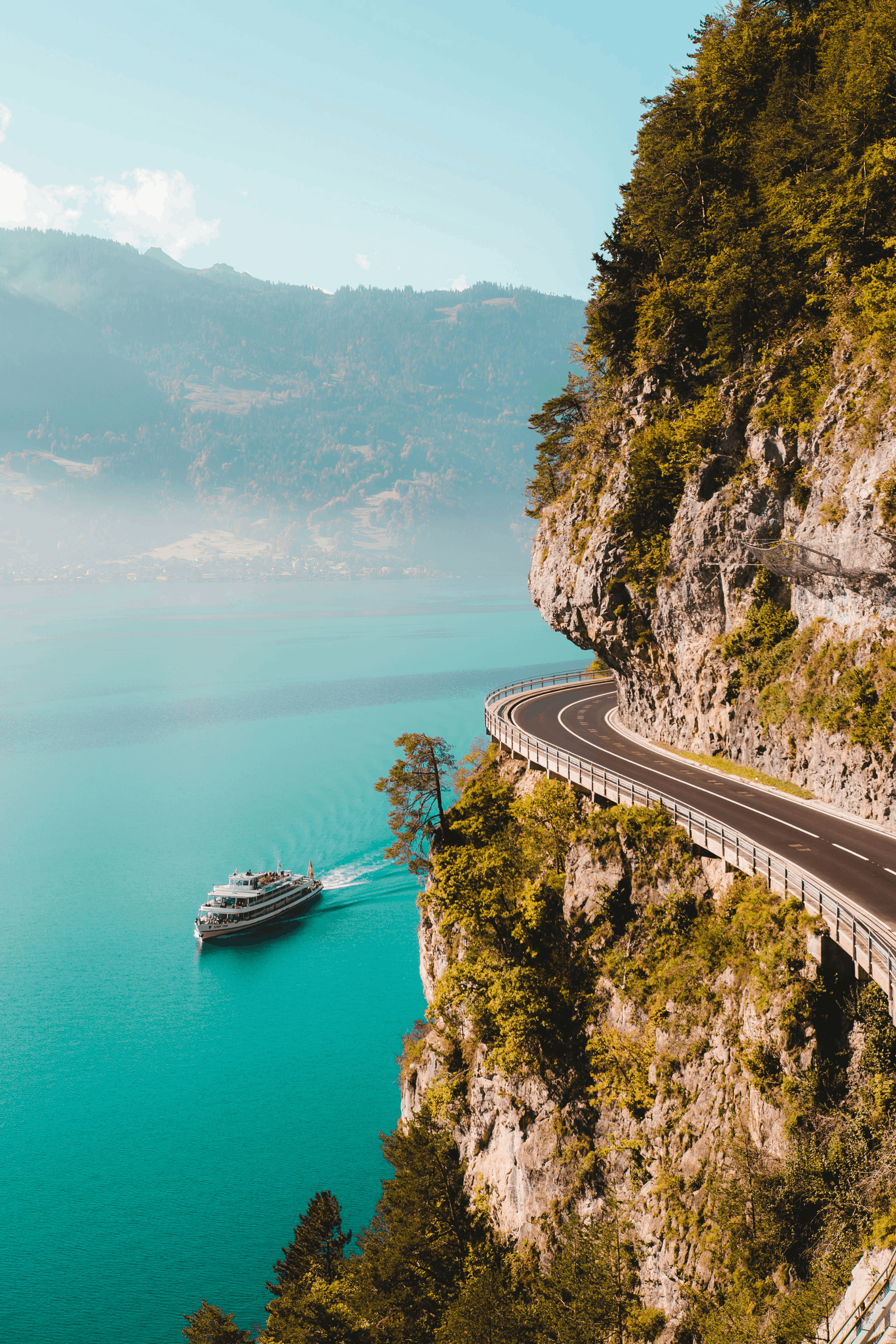 A long, winding road through rugged mountains with a white yacht visible in the distance on the water.