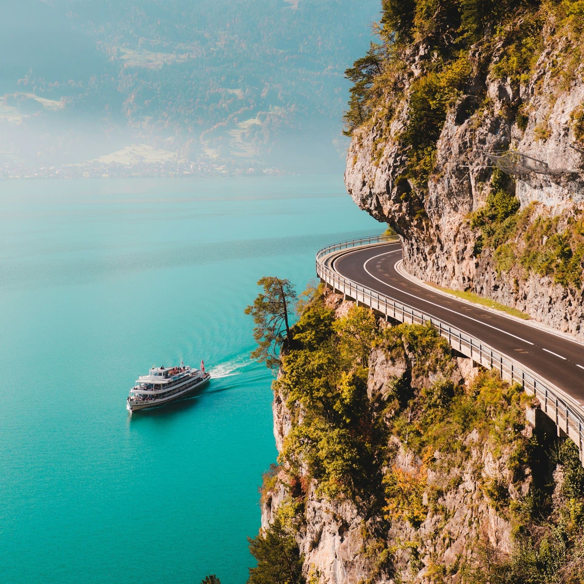 A long, winding road through rugged mountains with a white yacht visible in the distance on the water.