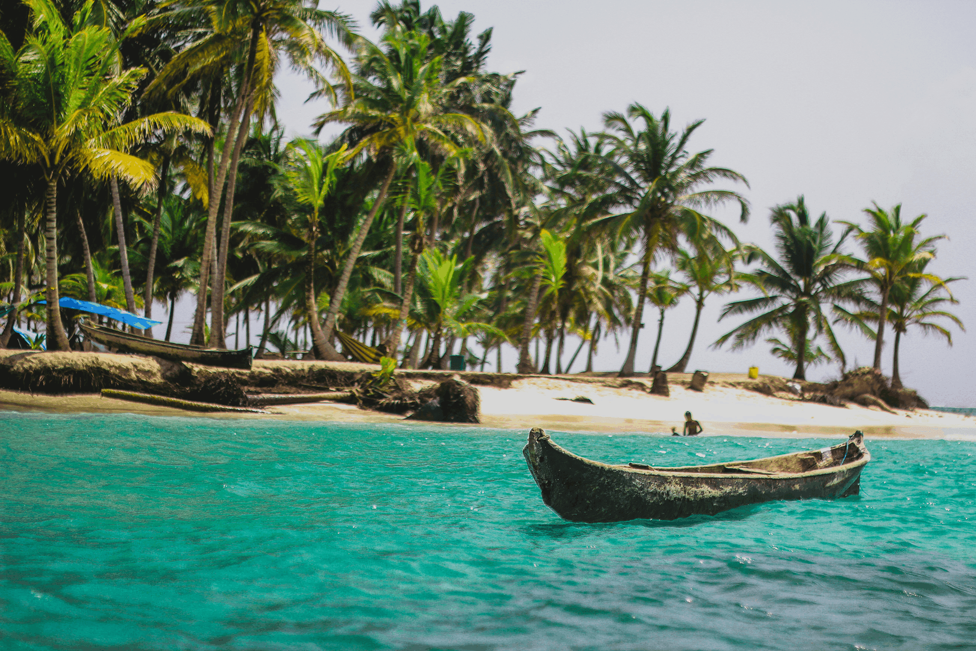 A rustic wooden boat in the foreground with people swimming and a palm-fringed island in the background.