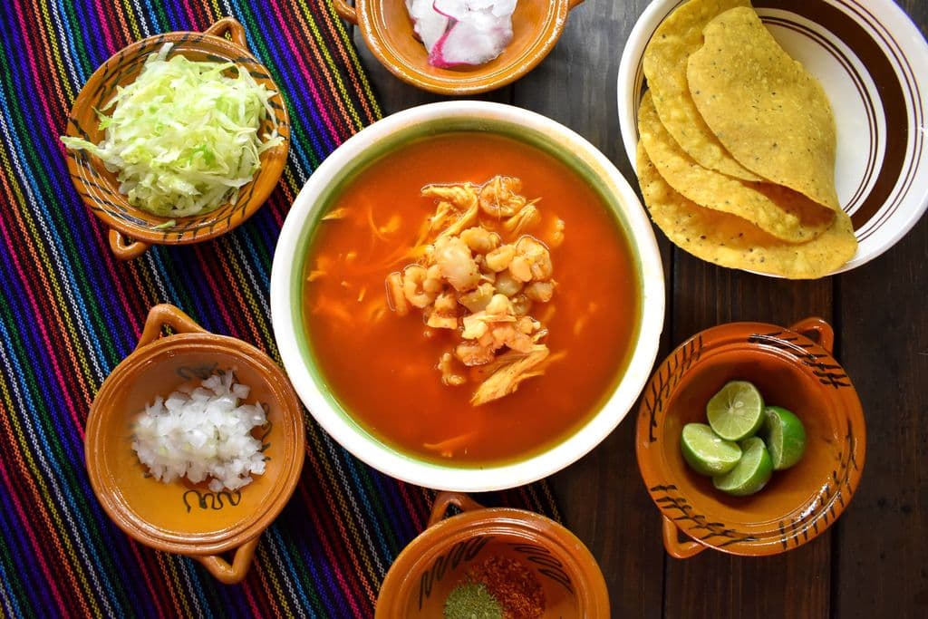 Bowl of traditional pozole with fresh garnishes, by Den Romi McRod